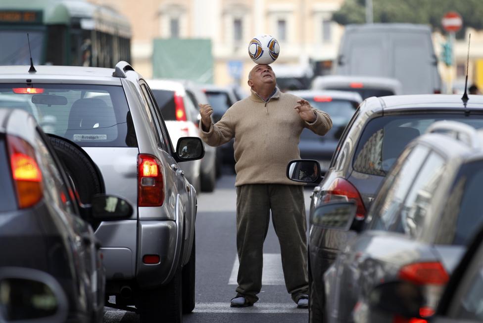 A street performer controls a soccer ball by his head as the cars are stopped in front of a crosslight in Rome